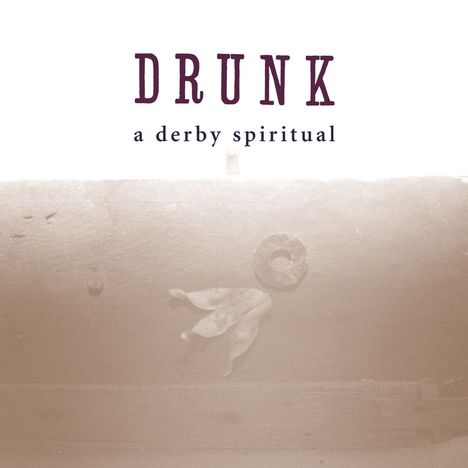 Text: "DRUNK a derby spiritual". Sepiafarbenes Foto mit einem Blatt und einer runden Metallscheibe auf Holz.