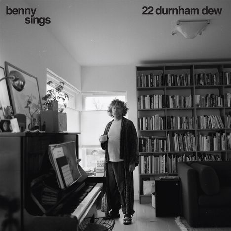 „benny sings“ und „22 durnham dew“. Ein Mann mit Tasse, im Morgenmantel, steht in einem Raum mit vielen Büchern und einem Klavier.