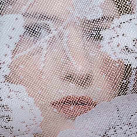 Ein Gesicht hinter durchscheinender weißer Spitze mit Blumenmuster, die Augen und Lippen leicht sichtbar.