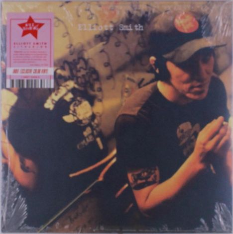 Text: "Elliott Smith". Der abgebildete Mensch trägt eine Mütze, sitzt in einem gedämpft beleuchteten Raum mit Kritzeleien.