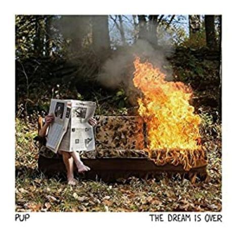 "PUP", "THE DREAM IS OVER". Ein Sofa brennt im Freien, daneben sitzt jemand mit einer Zeitung.