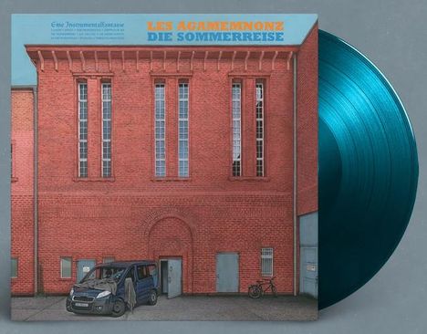 "LES AGAMEMNONZ DIE SOMMERREISE" in orange. Ein rotes Backsteingebäude mit hohen Fenstern, davor ein geparktes Auto.