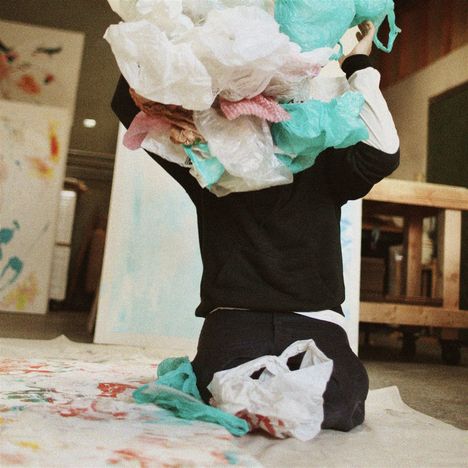 Eine Person kniet auf einem bemalten Boden, hält viele Plastiktüten über ihrem Kopf in einem Atelier.