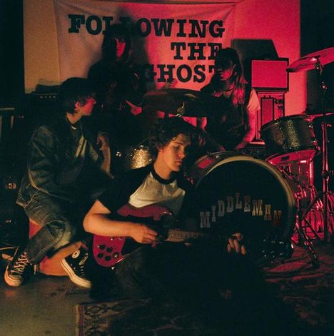 "FOLLOWING THE GHOST" auf Banner, "MIDDLEMAN" auf der Trommel. Vier Personen, Bandauftritt-Atmosphäre, schummriges Licht.