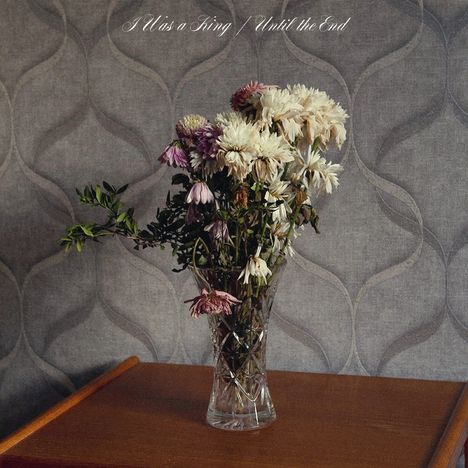 Text: „I Was a King / Until the End“. Ein Glasvase mit verwelkten Blumen steht auf einem Holzregal vor gemusterter Tapete.