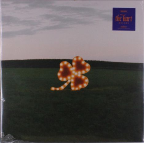 Text: "the hart deluxe." Ein beleuchtetes Kleeblatt auf einem Feld, im Hintergrund ist ein bewölkter Himmel zu sehen.