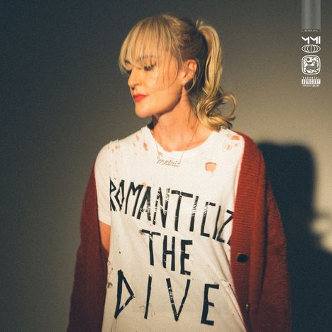 Der Text „ROMANTICIZE THE DIVE“ steht auf einem zerrissenen T-Shirt getragen von einer Frau mit roter Strickjacke und blonden Haaren.