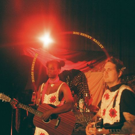 Texte: "BIG BENNY BAILEY", "BUSINESS SPICE". Zwei Musiker mit Gitarren, rotes Licht im Hintergrund, Kleidung mit Muster.
