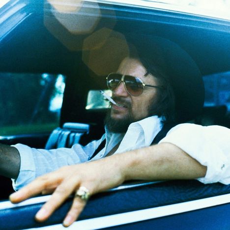 Ein Mann mit Hut und Sonnenbrille lehnt lässig im Auto, eine Zigarette im Mund.