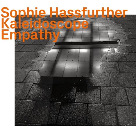 „Sophie Hassfurther Kaleidoscope Empathy“ in orange, über einem reflektierenden Pflastersteinboden mit Wasserlachen.