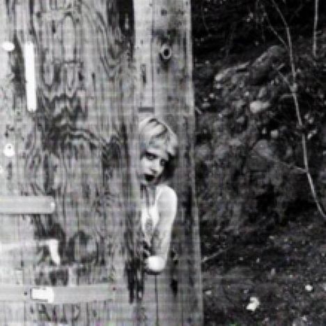 Ein schwarz-weißes Foto zeigt eine Person, die hinter einem großen Holzbalken hervorschaut, in einer Waldumgebung.