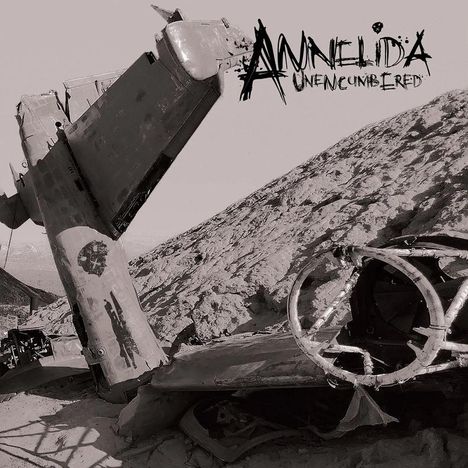 Text: "ANNELIDA" und "UNENCUMBERED". Ein zerbrochenes Flugzeugwrack in einer wüstenartigen Landschaft.