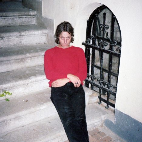 Es sind keine Texte vorhanden. Eine Person mit rotem Pullover sitzt auf Steinstufen neben einem kleinen, vergitterten Fenster.