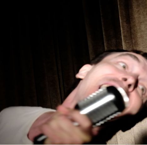 Ein Mann singt leidenschaftlich in ein Mikrofon vor einem dunklen Vorhang. Sein Gesicht ist dramatisch nach vorn geneigt.