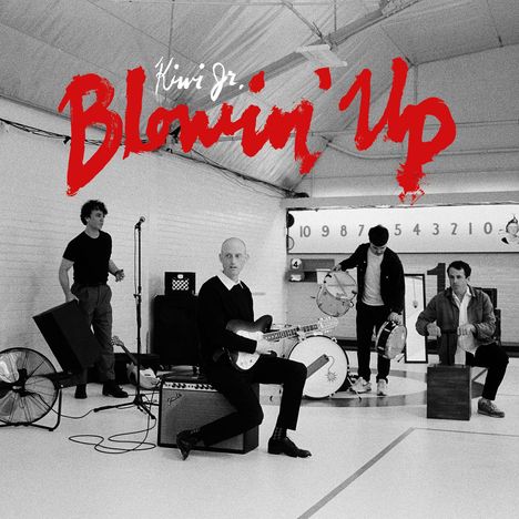 Text: "Kiwi Jr. Blowin' Up". Vier Männer in einem Raum mit Instrumenten. Einer spielt Gitarre, andere halten Percussion.