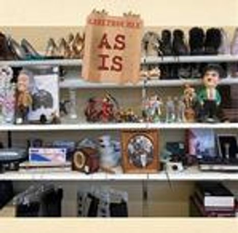 Der Text "AS IS" steht auf einem Schild. Darunter Schuhe, Figuren und diverse Gegenstände auf Regalen.