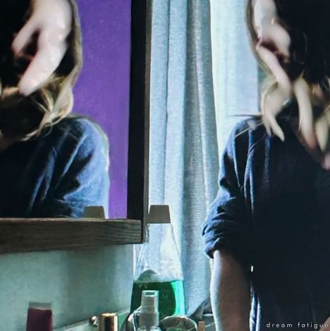 Text: "dream fatigue". Eine Person spiegelt sich in einem Badezimmer, umgeben von Flaschen und Kosmetikprodukten.