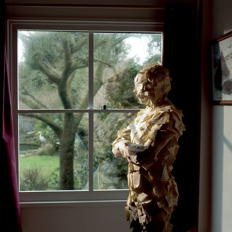 Ein Mensch, bedeckt mit vielen handschriftlich beschrifteten Notizzetteln, steht mit verschränkten Armen vor einem Fenster, mit Blick auf einen Garten.