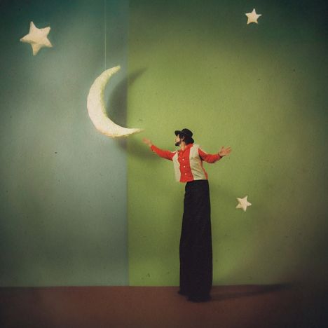 Ein Mensch auf Stelzen trägt Hut und farbenfrohe Kleidung, berührt einen Mond vor blauer und grüner Wand, umgeben von Sternen.