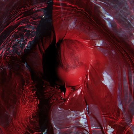Eine Person ist in rotes Tuch gehüllt, das von Wasser umgeben ist. Das Lichtspiel erzeugt einen dramatischen Effekt.