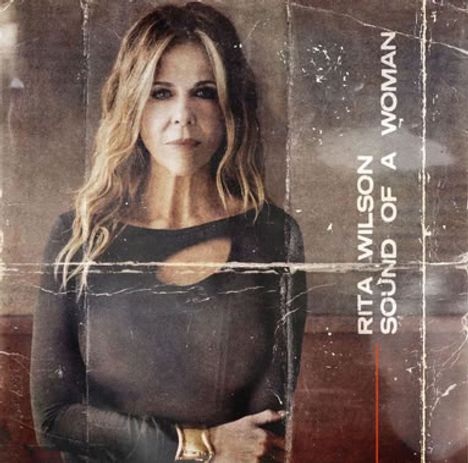 "RITA WILSON SOUND OF A WOMAN." Frau mit langen Haaren in einem dunklen Outfit vor einem abgenutzten Hintergrund.
