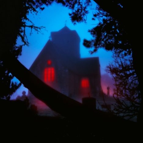 Eine mystische Kirche mit roten Fenstern, umgeben von Nebel und dunklen Bäumen im Vordergrund.