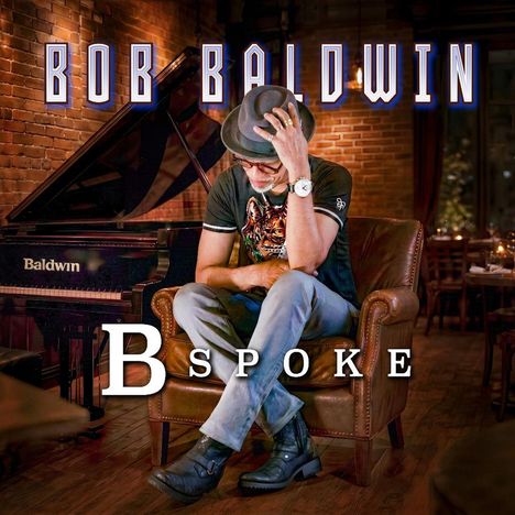 Oben steht "BOB BALDWIN", darunter "BESPOKE". Mann auf Sessel in Bar, Hut, Uhr, Flügel im Hintergrund.