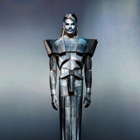 Keine Texte vorhanden. Eine metallisch aussehende Figur mit kantigen Schultern und futuristischem Aussehen steht frontal.