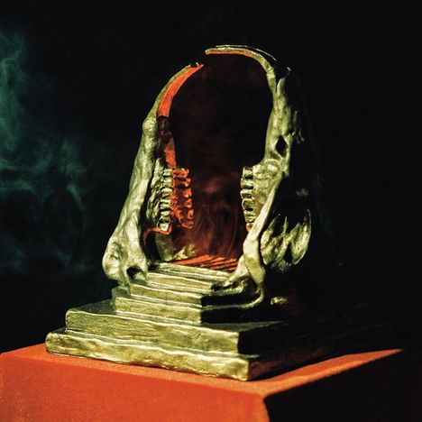 Eine dunkle, atmosphärische Skulptur, die wie ein geöffneter, organischer Torbogen mit angedeuteten Zähnen aussieht, auf einem Sockel.