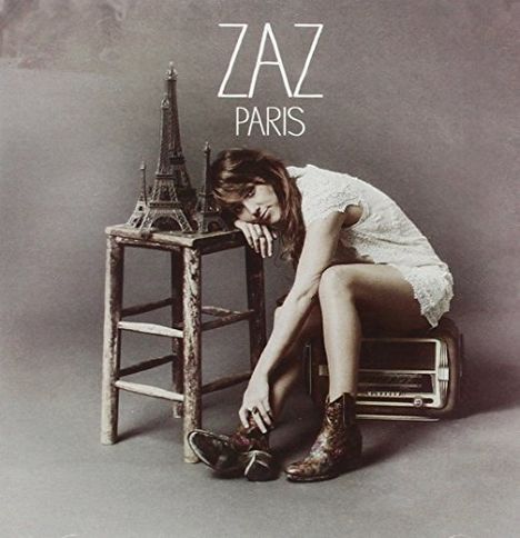 Text: "ZAZ PARIS". Eine Frau in einem weißen Kleid lehnt an einem Stuhl mit einem Eiffelturm-Modell und einem radioartigen Gegenstand.