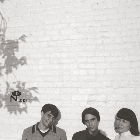 "No 233" links unten. Drei Menschen vor heller Ziegelwand. Schatten von Blättern fallen darauf.