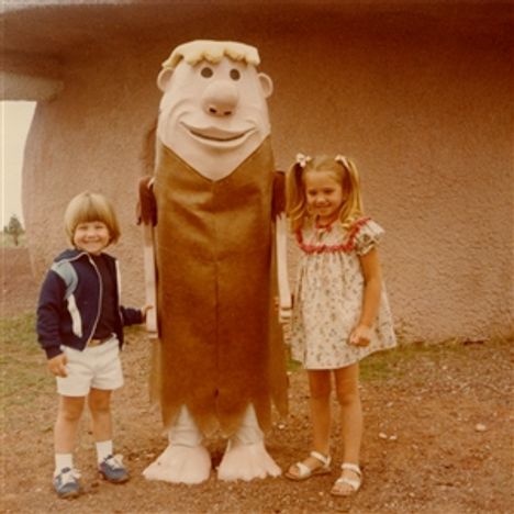 Zwei Kinder posieren fröhlich mit einer Figur im Höhlenmenschenkostüm, das große Augen und breites Lächeln hat.