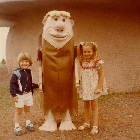 Ich kann darauf keine Texte erkennen. Zwei Kinder stehen lächelnd neben einer großen Cartoon-Figur mit großer Nase.