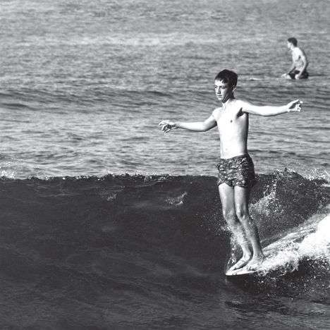 Zwei Jungen surfen auf kleinen Wellen. Einer steht auf dem Surfbrett, der andere sitzt im Hintergrund im Wasser.