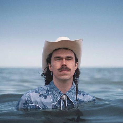 Ein Mann mit Cowboyhut steht bis zur Brust im Wasser, trägt ein blaues Hemd mit floralen Mustern.