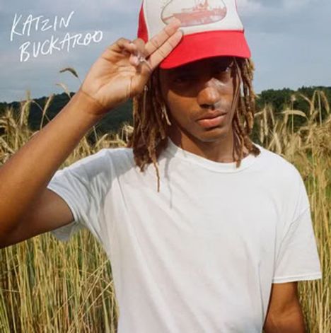 „KAIZIN BUCKAROO“ steht links oben. Eine Person in weißem T-Shirt und rotem Cap steht in einem Feld.