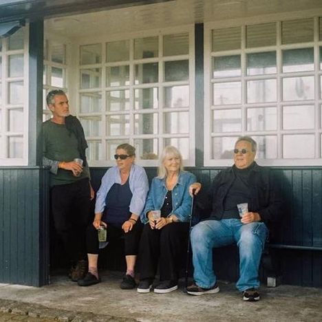 Vier Menschen sitzen entspannt auf einer Bank in einem überdachten Bereich mit vielen Fenstern, teilweise lächelnd.