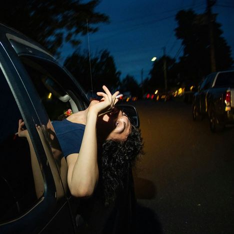 Person lehnt aus Autofenster, raucht eine Zigarette. Straße im Hintergrund, abends oder nachts.