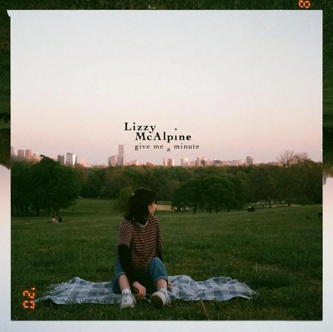 „Lizzy McAlpine – give me a minute“ über einem Park. Eine Person sitzt auf einer Decke mit Blick auf die Skyline.