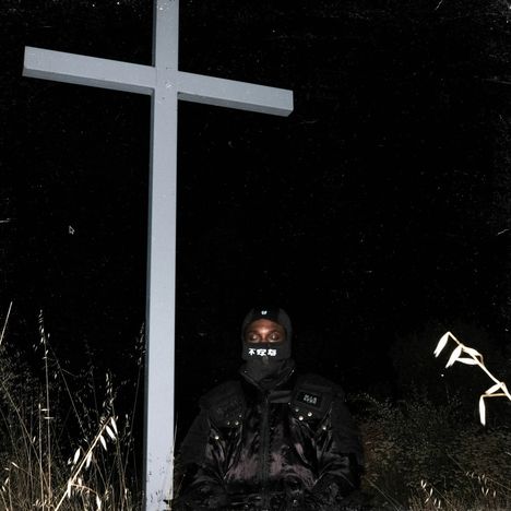 Jemand in schwarzer Kleidung vor einem hohen Kreuz. Sie tragen eine Maske mit weißen Schriftzeichen und stehen im Gras.