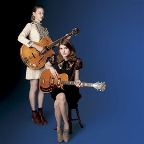 Zwei Frauen mit Gitarren auf blauem Hintergrund, eine sitzend, die andere stehend, beide in eleganter Kleidung.