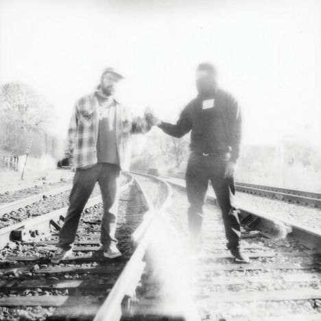 Zwei Menschen stehen auf Bahngleisen und geben sich die Hand. Es wirkt wie ein nostalgisches, verblasstes Foto.