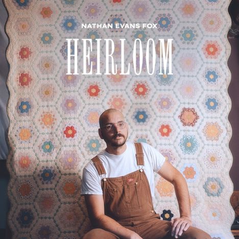 Text: "NATHAN EVANS FOX HEIRLOOM". Ein Mann sitzt vor einer bunten Patchwork-Decke, trägt Brille und braune Latzhose.