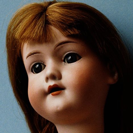 Keine erkennbaren Texte. Großaufnahme eines Puppenkopfes mit blauen Augen und braunem Haar vor blauem Hintergrund.