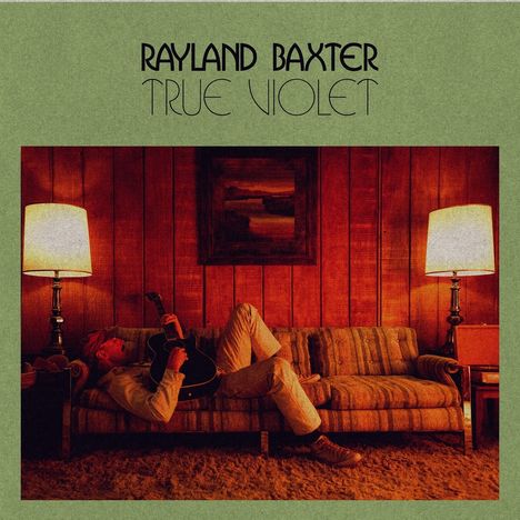 "Rayland Baxter True Violet." Eine Person liegt mit Gitarre auf einem Sofa zwischen zwei Lampen, Holzwand und Gemälde dahinter.
