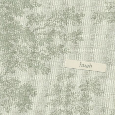 „Hush“ steht auf einem kleinen Etikett. Der Hintergrund hat ein dezentes, florales Muster in Grau auf beigem Stoff.