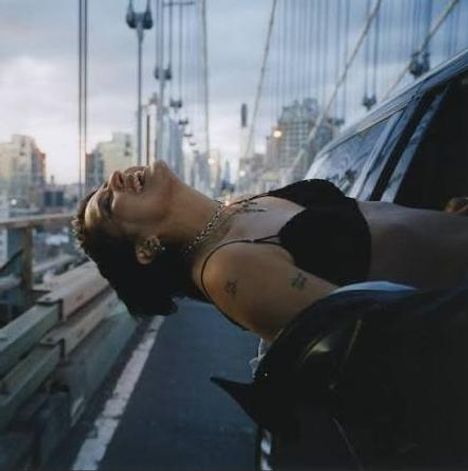 Eine Person lehnt sich lachend aus einem Auto auf einer Brücke. Die Stadt ist im Hintergrund bei Dämmerung zu sehen.