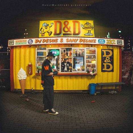 Ein gelber Imbissstand mit "D&D" und "DJ DESUE & SAMY DELUXE" Text. Außen eine große Eiscreme-Attrappe.