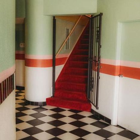 Kein Text. Eine Treppe mit rotem Teppich führt nach oben, umgeben von grünen Wänden und schwarz-weißem Fliesenboden.