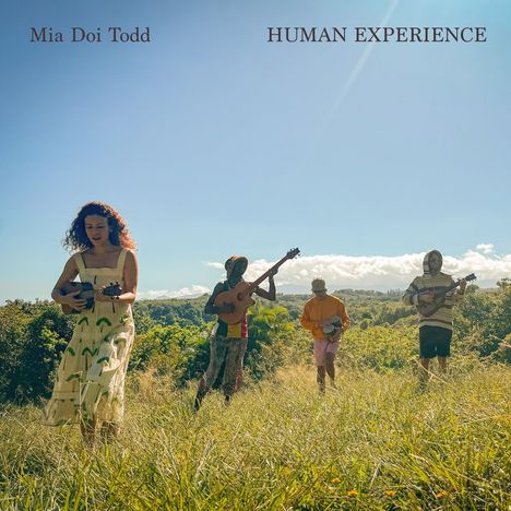 Text: "Mia Doi Todd HUMAN EXPERIENCE". Vier Personen spielen Instrumente auf einer Wiese an einem sonnigen Tag.